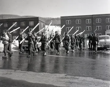 group of skiers cross a road in front of two brick buildings