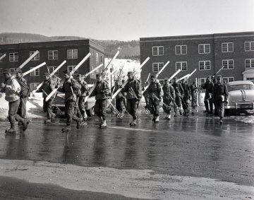 group of skiers cross a road in front of two brick buildings
