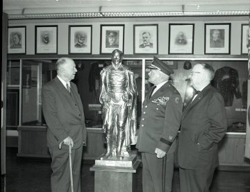 three men, one with cane, one in military uniform, gaze at a statue