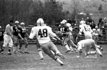 football player in dark uniform runs with ball between three defenders in white, two players in dark uniform look on