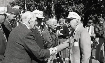 two older men shaking hands, man at right wearing a baseball cap