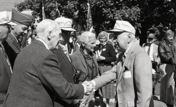 two older men shaking hands, man at right wearing a baseball cap
