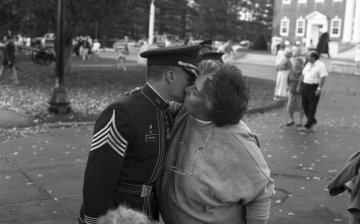 Cadet in uniform embraces woman, people in background walking toward them
