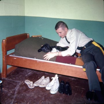 cadet in dress shirt and uniform pants petting dog on a bunk bed