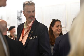 man with glasses on forehead and wearing a suit, name badge and lanyard glances in a downward direction, with a woman directly behind.