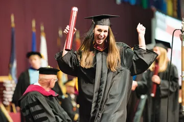 Woman in graduation gown and cap, in an arms-raised celebratory gesture, holding up her diploma (in a cylinder) as she walks across the graduation ceremony stage. Photo of Clara leister '16.