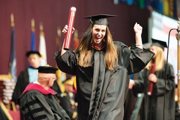 Woman in graduation gown and cap, in an arms-raised celebratory gesture, holding up her diploma (in a cylinder) as she walks across the graduation ceremony stage. Photo of Clara leister '16.