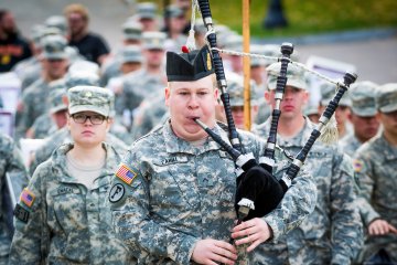 A large group of Norwich cadets in military camo fatigue uniform trail a man playing bagpipes. Photo from Norwich University's 2015 Legacy March.
