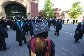 An individual in graduation gown and cap, with the cap reading "Enjoy Life", appears from behind, among a group of fellow graduates. Photo of NU CGCS master's candidates preparing to line up for their graduation ceremony.
