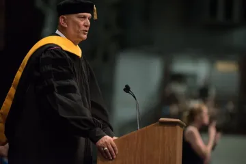 A man in black graduation gown and yellow sash speaks at a lectern. Photo of Gen. Raymond Odierno’s 2016 Graduation Address, 05.14.16.