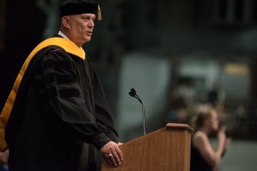 A man in black graduation gown and yellow sash speaks at a lectern. Photo of Gen. Raymond Odierno’s 2016 Graduation Address, 05.14.16.