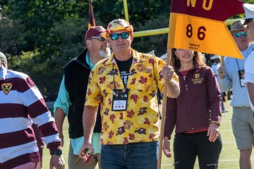 man in sunglasses, bright yellow floral shirt and boat cap, holding a guidon flag with NU-86 written on it. Several individuals follow him. Photo of guidon holder Phil Baumann '86, M'14 & P'19.
