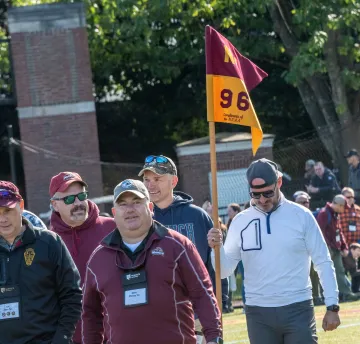 Man in backwards baseball cap and white long-sleeve shirt carrying guidon flag with "N-96" written on it. Several other men walk beside him. Photo from Norwich University 2022 Homecoming Alumni Parade.
