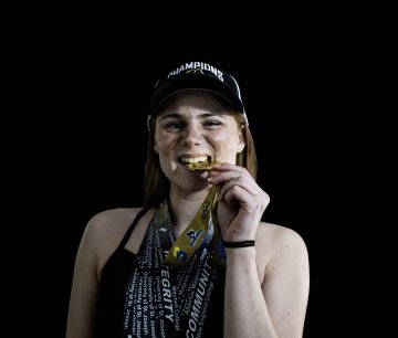 Person wearing a champions hat holds several medals on ribbons against a black background.