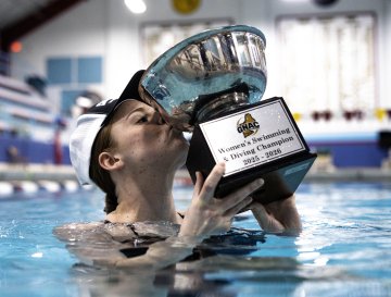 Person in a pool lifts a large trophy labeled “Women’s Swimming & Diving Champion 2025–2026.”