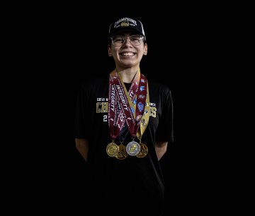 Person in a black shirt and hat stands against a black background wearing several medals on wide ribbons.