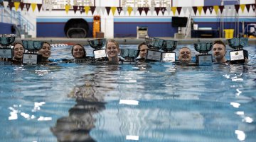 Swimmers in a pool hold several trophies above the water near the pool’s edge.