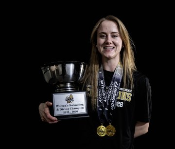 Person in a black shirt holds a large trophy labeled “Women’s Swimming & Diving Champion 2025–2026” with medals hanging from their neck.