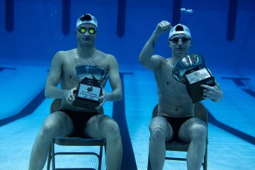 Two swimmers sit on chairs underwater holding large championship trophies.