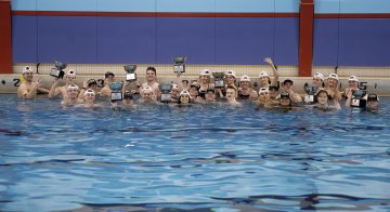 A large group of swimmers in a pool hold up trophies while gathered near the pool’s edge.