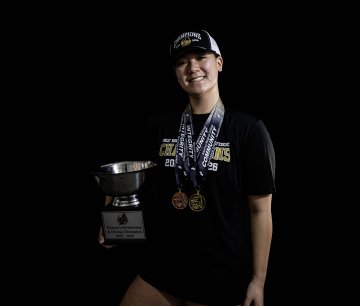Person in a black shirt and hat holds a large trophy and wears several medals against a black background.