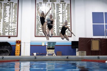 Four swimmers in black suits jump into a pool holding a trophy with record boards hanging on the wall behind them.