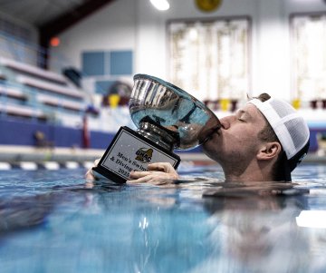 Person in a pool lifts a large trophy labeled “Men’s Swimming and Diving Champion 2025–2026.”