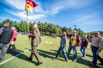 Man in olive-colored military themed jacket and cap, between two individuals in front and four behind, carries a guidon flag reading "NU 86".