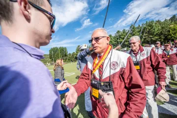 Man in red & white jacket wearing a medal shakes hands with a younger individual. Photo of Claudius Colombo '71 at Norwich Univ. Homecoming 2021. 