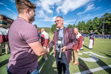 Man in burgundy sport jacket holding a long lens camera shakes hands with a younger individual. Photo of Don Hirsch Jr. '71 at Norwich Univ. Homecoming 2021. 