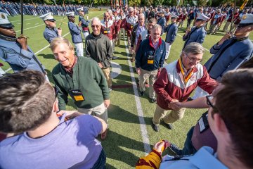Two men, on left in green henley shirt, on right, in a red & white jacket, approaching other individuals to shake hands. 