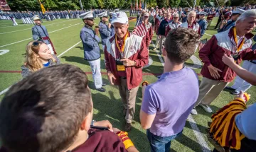 Man in red & white jacket approaches three younger individuals holding his phone. Photo of Greg May '71 at Norwich Univ. Homecoming 2021. 