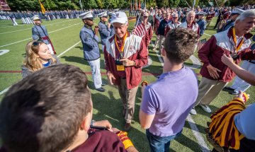 Man in red & white jacket approaches three younger individuals holding his phone. Photo of Greg May '71 at Norwich Univ. Homecoming 2021. 