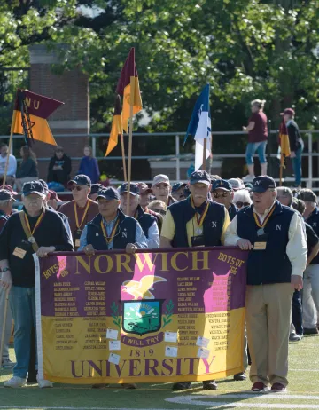 group of men heading onto a football field, front of group holding a maroon and gold banner stating Norwich University. Photo from Norwich Univ. Homecoming 2019. 