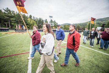 Two women and two men walk across a football field. Woman leading the group in holding a guidon flag reading "NU 91".