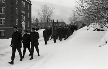 Black and white photo of a group of Norwich cadets in dark outerwear marching in the snow by a brick building.