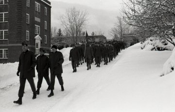 Black and white photo of a group of Norwich cadets in dark outerwear marching in the snow by a brick building.