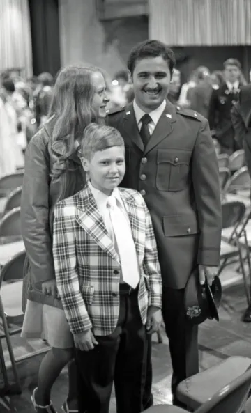 Black and white photo of a boy in plaid suit jacket and tie, woman in light colored leather jacket, and man in formal military suit. Part of Commissioning Ceremonies at Norwich University, 1976.