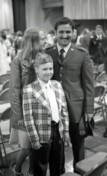 Black and white photo of a boy in plaid suit jacket and tie, woman in light colored leather jacket, and man in formal military suit. Part of Commissioning Ceremonies at Norwich University, 1976.