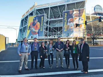A line of individuals in business casual clothing stand before the wall of a stadium with larger banners for the Denver Broncos and Carolina Panthers. Photo of the cybersecurity detail of Norwich students including Josh Hartley '16 at far left.