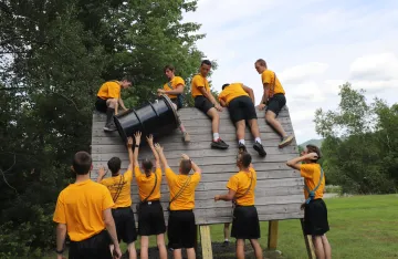 FLC campers working together to lift a large barrel over a wall as part of the challenge course.