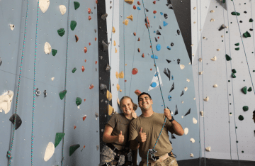 Two FLC campers smiling with thumbs up in front of rock climbing wall.
