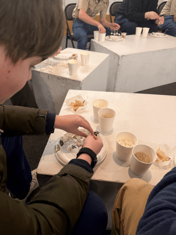 People sit at white tables with cups, toothpicks, snacks, and paper plates during an indoor activity.