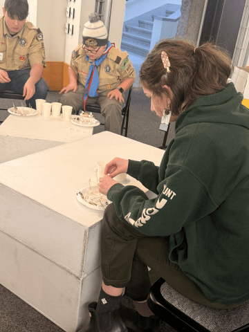 People sit around white tables working with small items on paper plates and cups during an indoor activity.