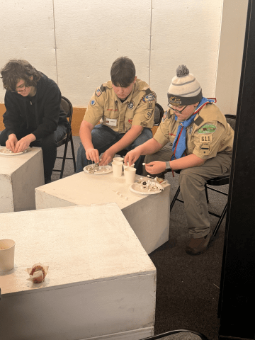 Three people sit at white tables working with small items on paper plates and cups during an indoor activity.