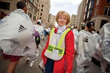 woman in yellow reflective vest and orange jackets stands as running race finishers wrapped in mylar blankets move in opposite direction behind her. Michelle Kanavos.