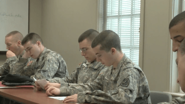 several Norwich cadets in military fatigues sit together at a desk. Video still from: "Thanks for making the Norwich Forever! campaign a success."