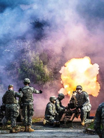 Five Norwich Cadet artillery battery cadets in uniform stand and kneel before a cannon firing, with a bright yellow plume of flame coming from the cannon. Photo from Veterans Day Ceremonies 11/10/2015.
