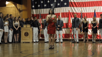 A woman in a red dress on a basketball court twirling a rifle before a group of students. Video still from: " Spring: Norwich embraces the warm weather"
