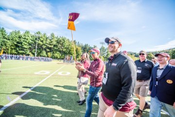 A group men walk to approach the 50-yard line on a football field, being led by a man in red visor and jacket, with another woman by his right side holding a guidon flag reading "NU 91". Photo of Edward Sullivan, John  Alexander, William McCollough, all  '91 (among others) at Norwich Univ. Homecoming 2021. 